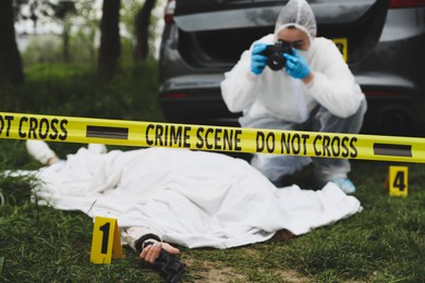 Photo of Criminologist taking photo of dead body at crime scene outdoors, focus on yellow tape