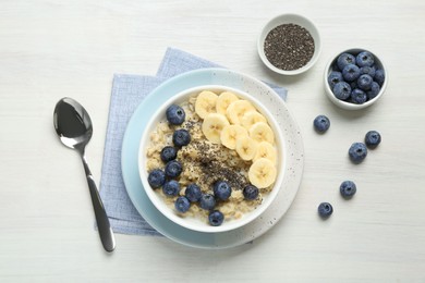 Tasty oatmeal with banana, blueberries and chia seeds served in bowl on white wooden table, flat lay