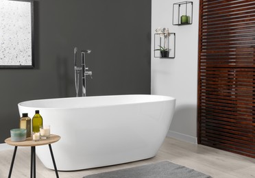 Photo of Stylish bathroom interior with ceramic tub and care products on coffee table