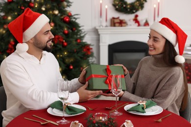 Beautiful young woman in Santa hat presenting Christmas gift to her boyfriend at served table in decorated room