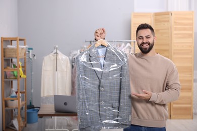 Photo of Dry-cleaning service. Happy man holding hanger with jacket in plastic bag indoors, space for text