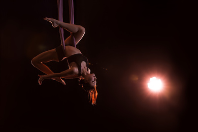 Young woman performing acrobatic element on aerial silk against dark background. Space for text