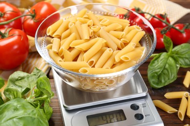 Photo of Kitchen scale with bowl of pasta among tomatoes and basil on wooden table, closeup