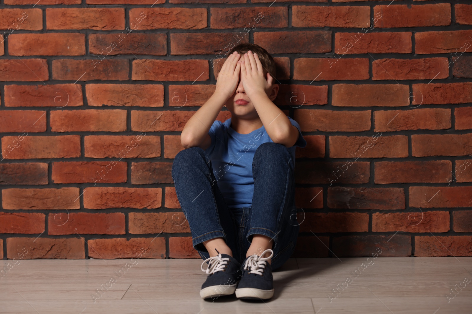 Photo of Child abuse. Upset boy sitting on floor near brick wall indoors