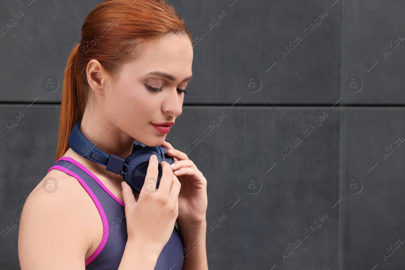 Photo of Beautiful woman in stylish gym clothes with headphones near dark grey wall on street, space for text