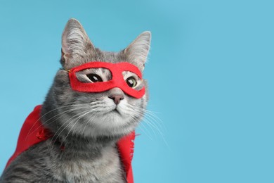 Photo of Adorable cat in red superhero cape and mask on light blue background, space for text