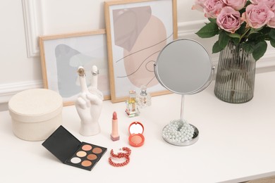 Mirror, cosmetic products, perfumes and vase with pink roses on white dressing table