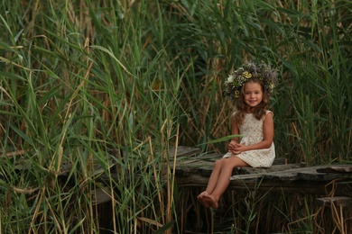 Photo of Cute little girl wearing wreath made of beautiful flowers on wooden bridge