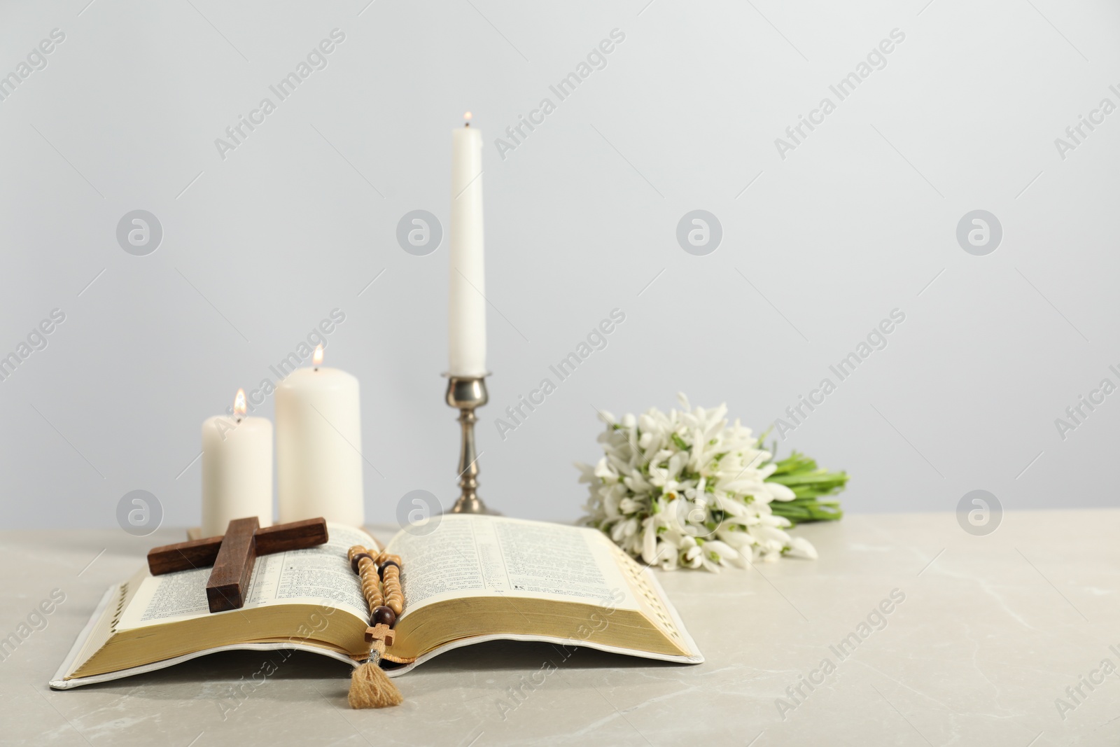 Photo of Church candles, wooden cross, rosary beads, Bible and flowers on light table. Space for text