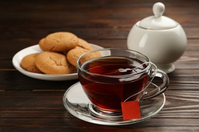 Photo of Tea bag in glass cup of hot water and cookies on wooden table