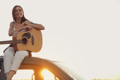Photo of Happy woman with guitar sitting on car roof outdoors at sunset. Summer trip