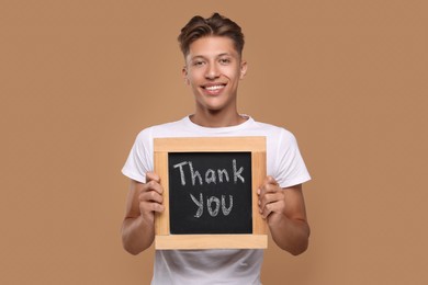 Photo of Happy man holding small chalkboard with phrase Thank You on brown background