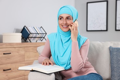 Muslim woman with book talking on smartphone on couch in room