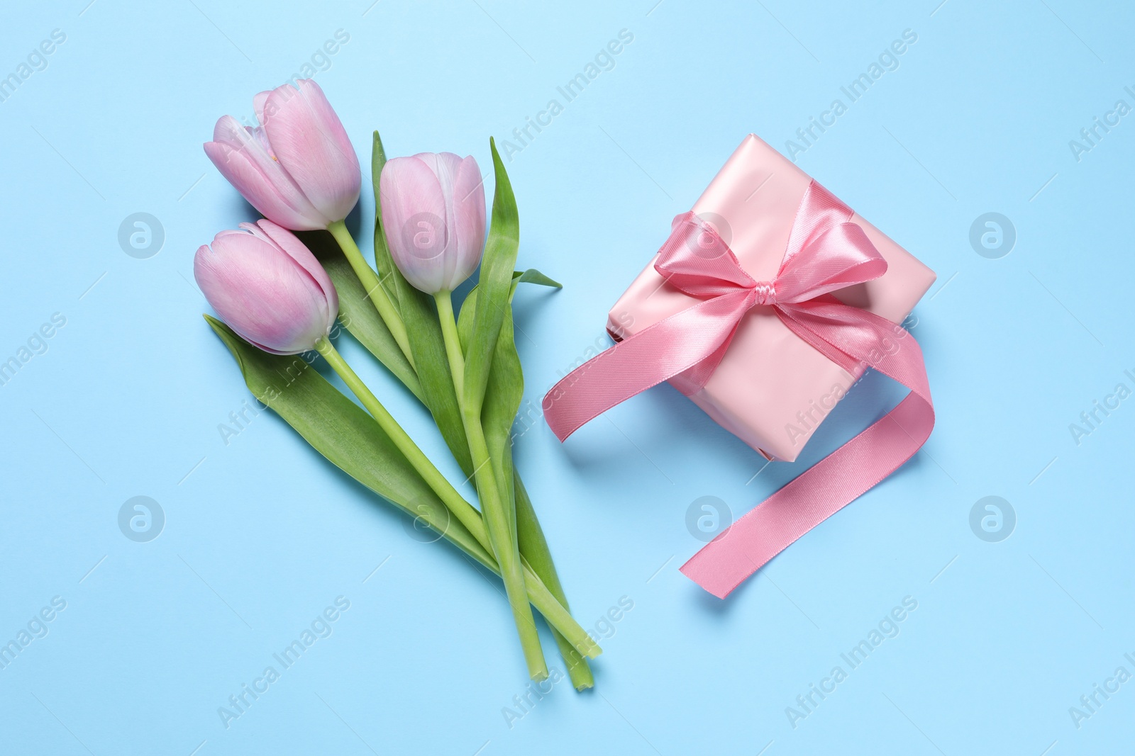 Photo of Beautiful gift box with bow and tulips on light blue background, flat lay