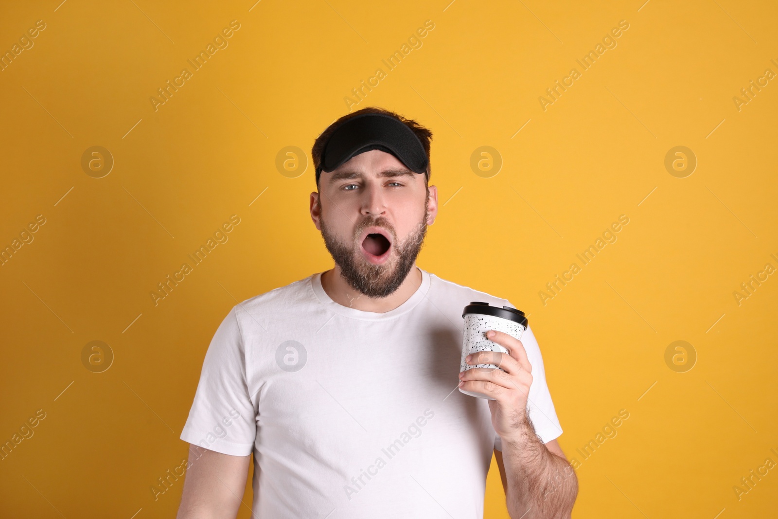 Photo of Tired young man with sleep mask and cup of coffee yawning on yellow background