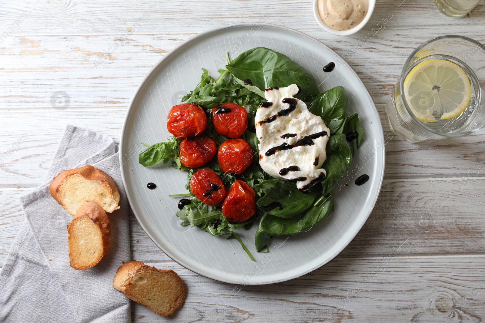 Photo of Delicious burrata cheese served with tomatoes, croutons, arugula and basil on white wooden table, flat lay