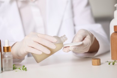 Dermatologist with bottle testing cosmetic product at white table indoors, selective focus