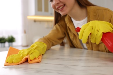 Photo of Woman with spray bottle and microfiber cloth cleaning white marble table in kitchen, closeup