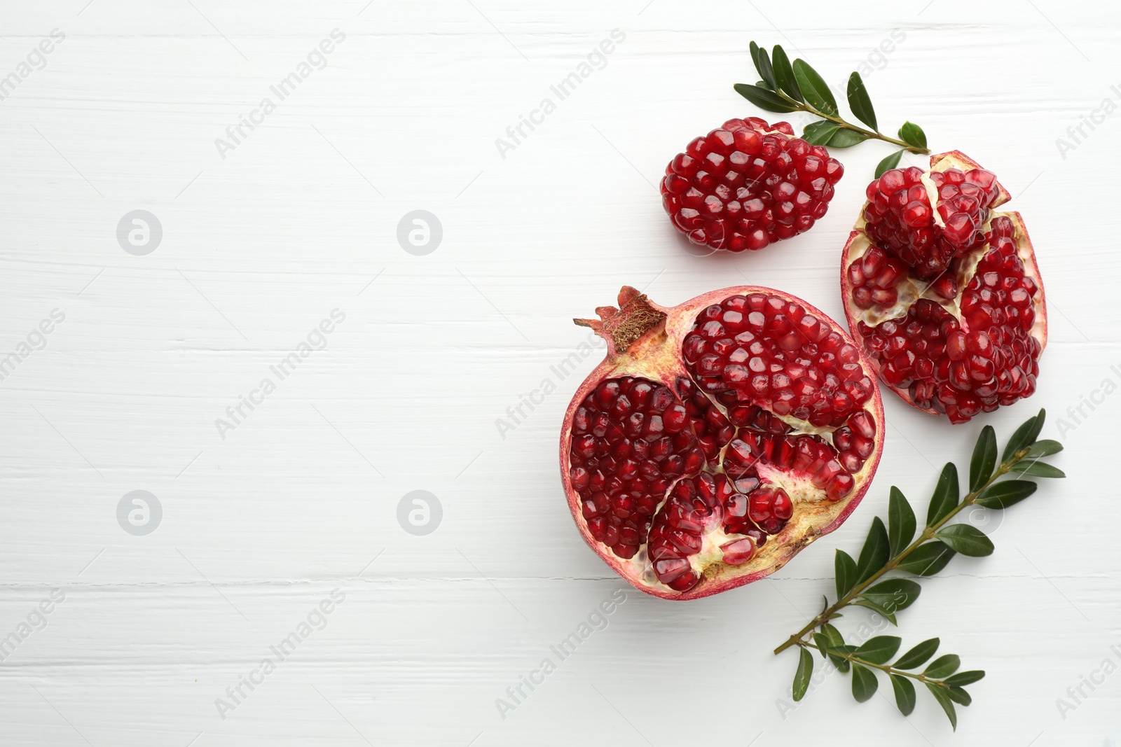 Photo of Pieces of fresh pomegranate and branches on white wooden table, flat lay. Space for text