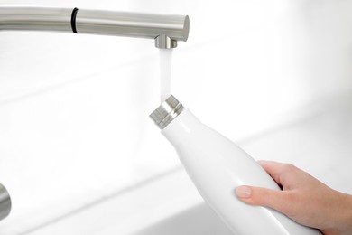 Photo of Woman pouring fresh water from tap into thermo bottle indoors, closeup