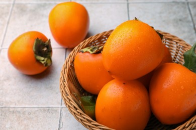 Photo of Delicious ripe juicy persimmons in wicker basket on tiled surface, closeup