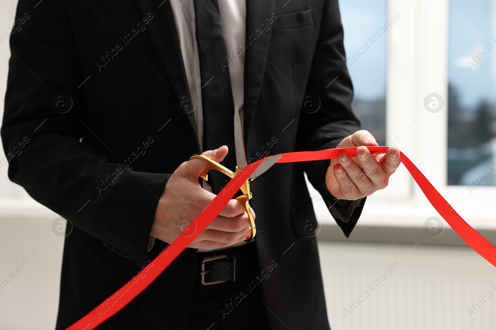 Photo of Man cutting red ribbon with scissors indoors, closeup