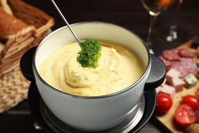 Photo of Dipping piece of broccoli into fondue pot with melted cheese at table with snacks, closeup