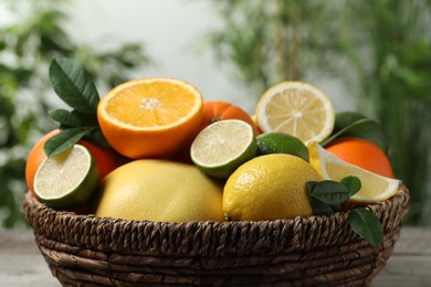 Different fresh citrus fruits and leaves in wicker basket against blurred background, closeup