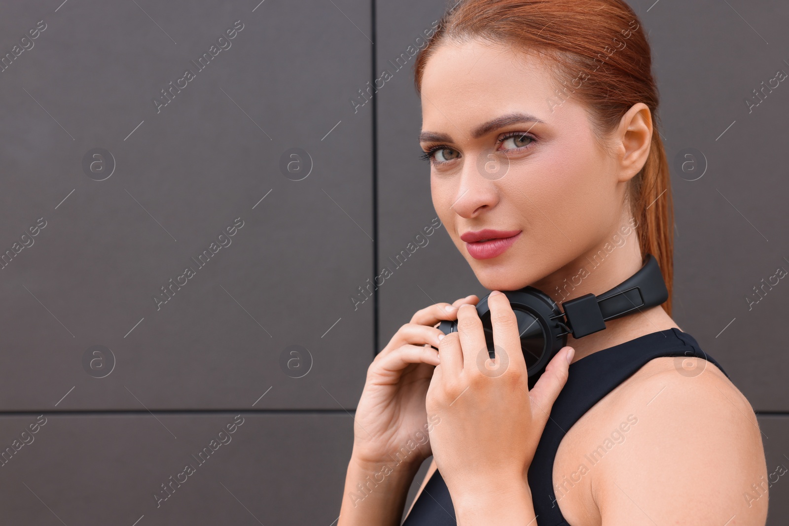 Photo of Beautiful woman in stylish gym clothes with headphones posing near dark grey wall on street, closeup. Space for text