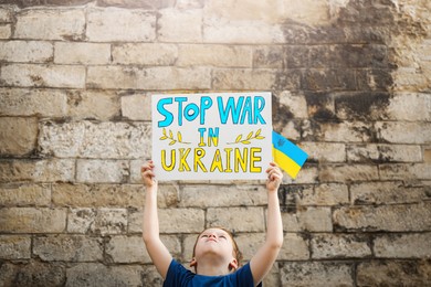Photo of Boy holding poster Stop War In Ukraine and national flag against brick wall outdoors