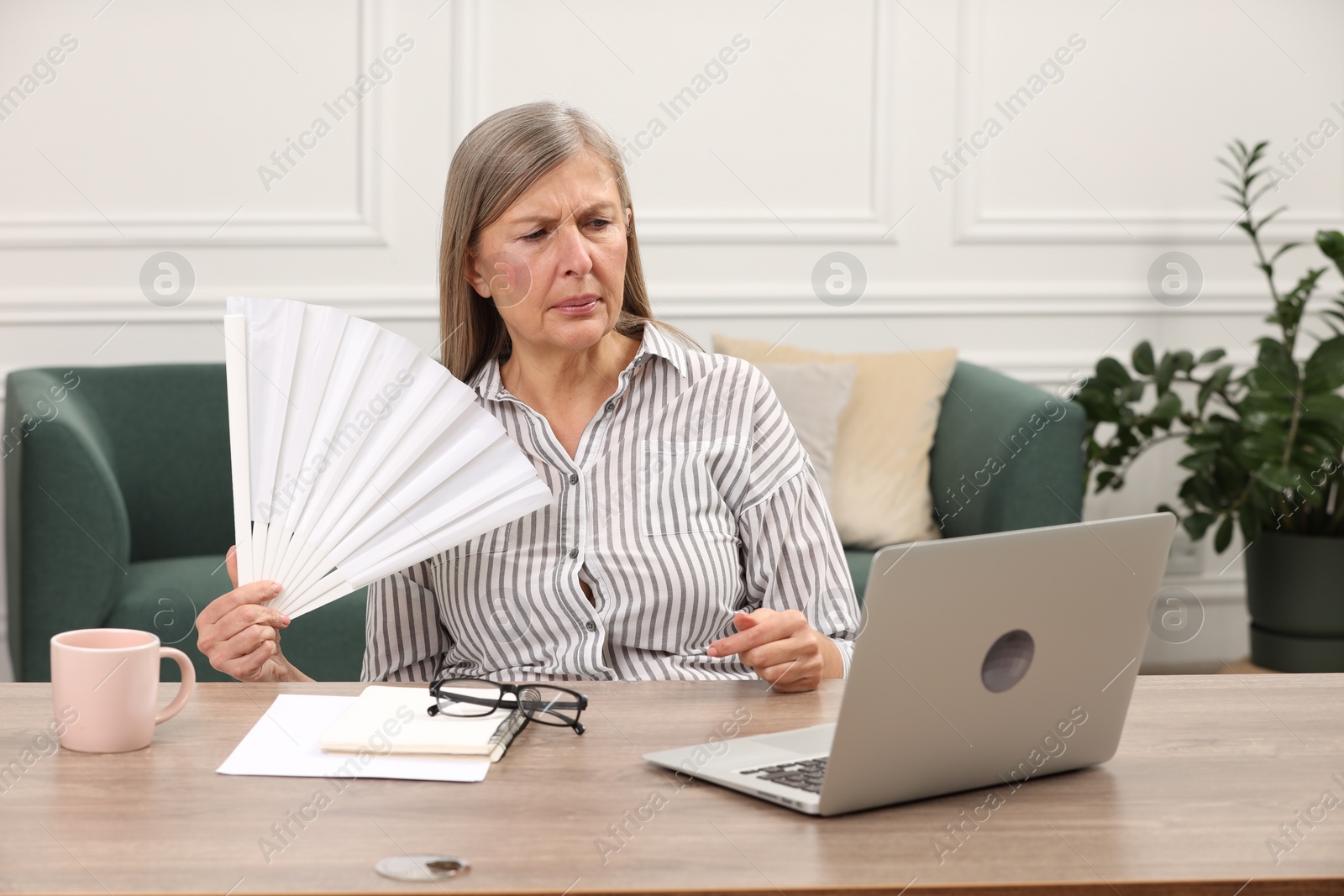 Photo of Menopause. Woman waving hand fan to cool herself during hot flash at wooden table indoors