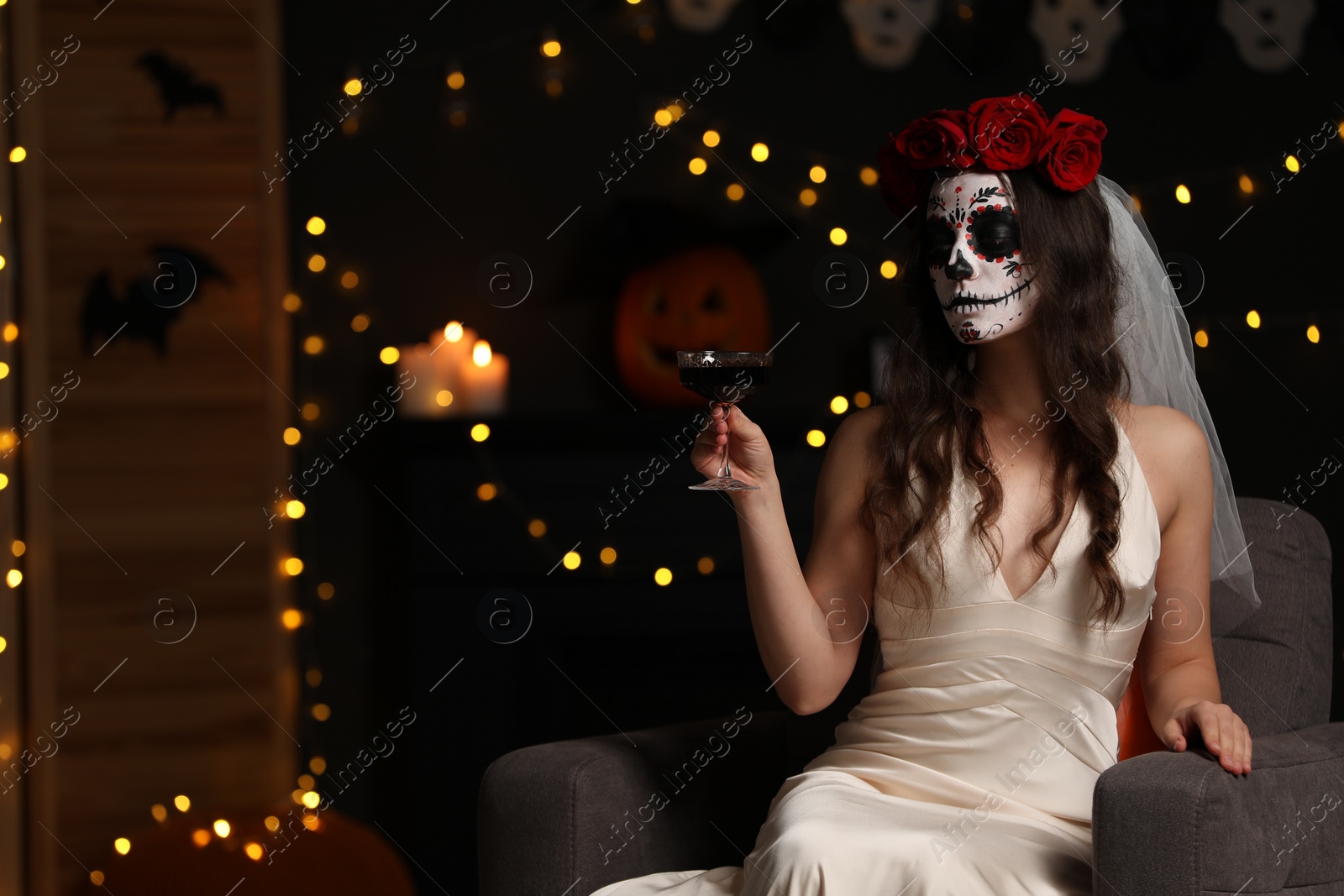 Photo of Young woman in scary bride costume with sugar skull makeup and glass of wine against blurred lights indoors, space for text. Halloween celebration