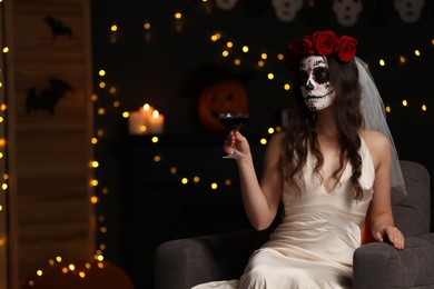 Photo of Young woman in scary bride costume with sugar skull makeup and glass of wine against blurred lights indoors, space for text. Halloween celebration