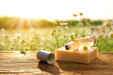 Photo of Roller bottle with chamomile essential oil and soap bar on wooden table in field