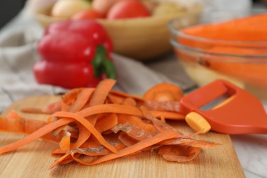 Photo of Wooden board with carrot peels and peeler on white table, closeup