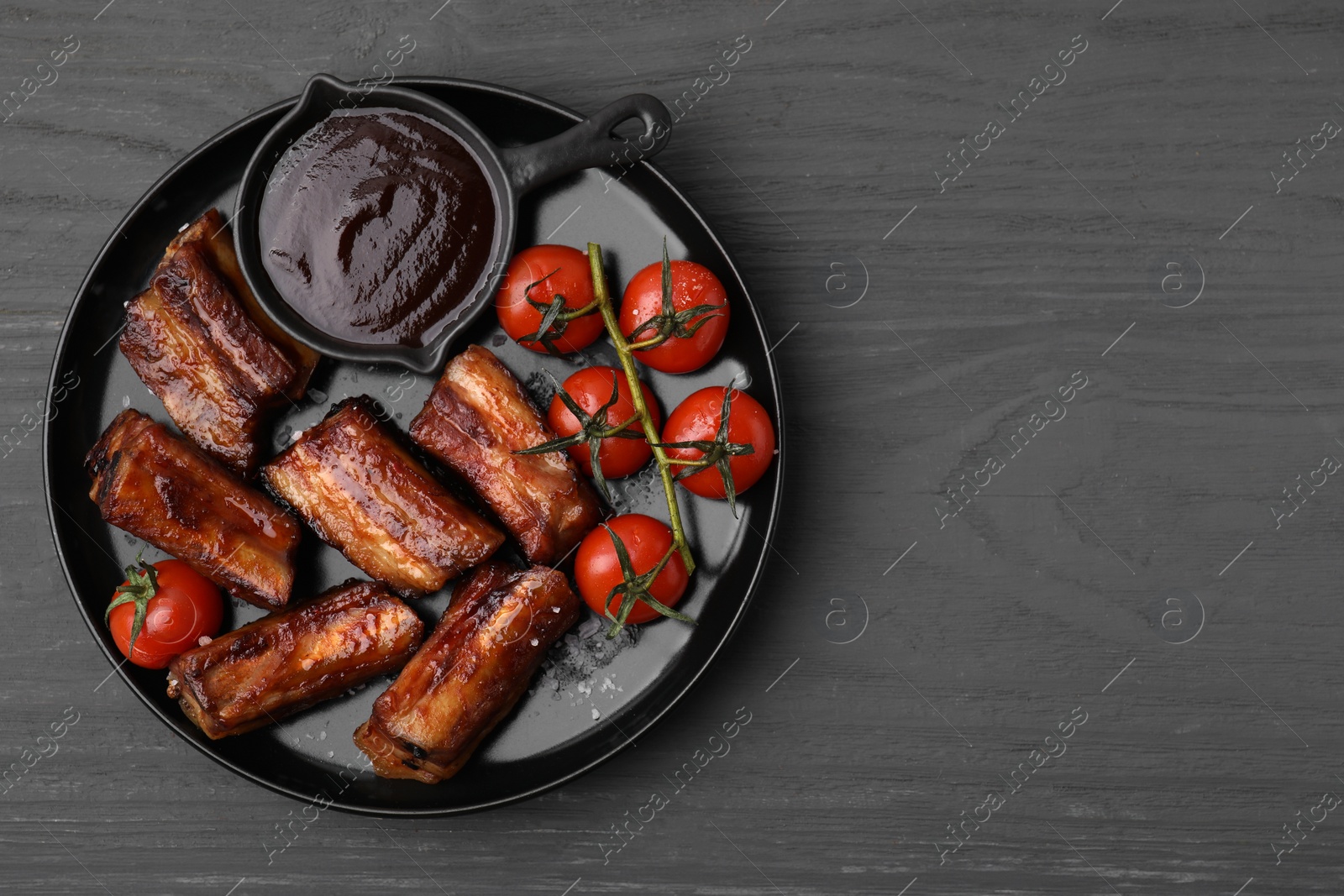 Photo of Tasty roasted pork ribs served with sauce and tomatoes on grey wooden table, top view. Space for text