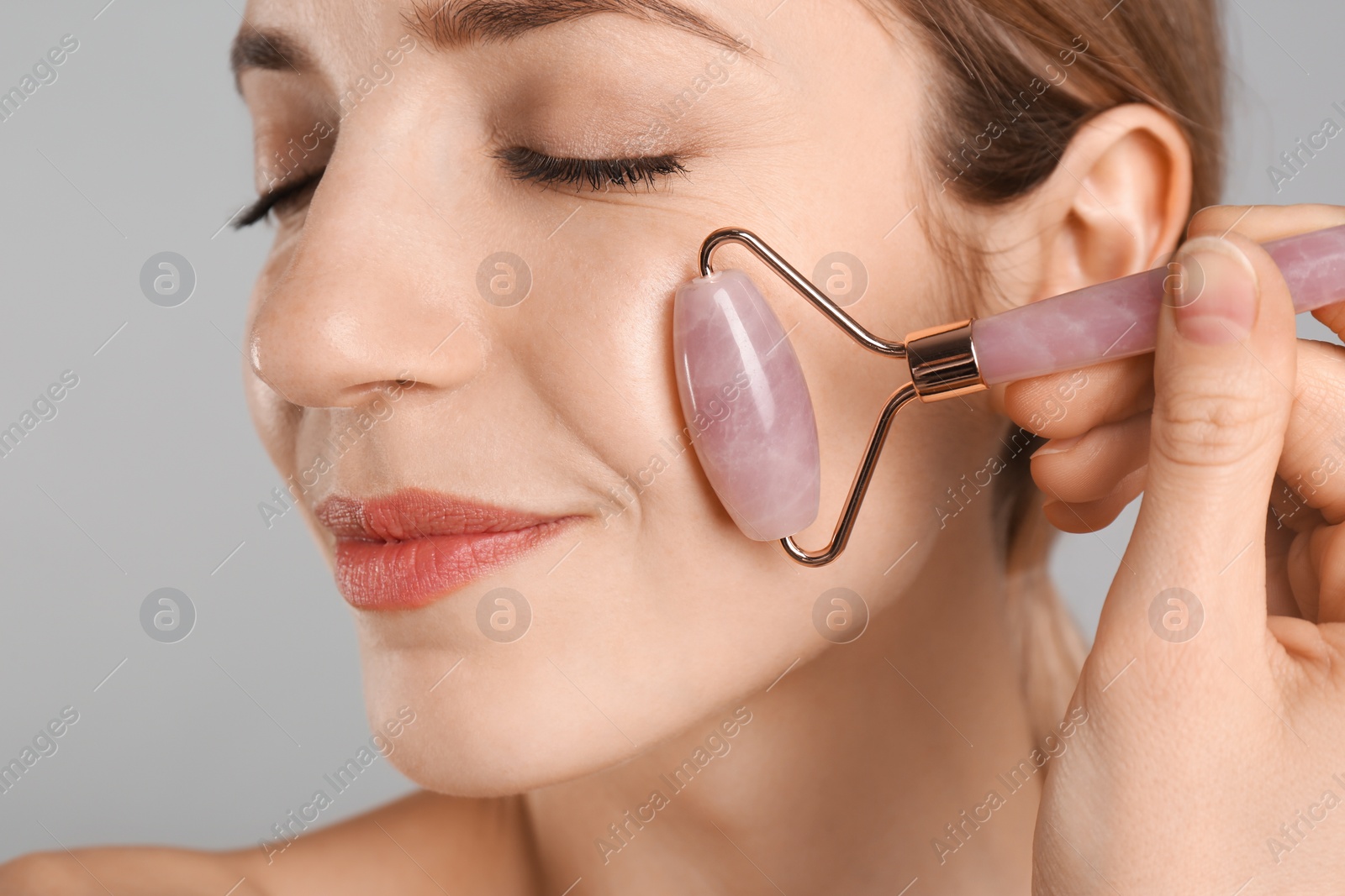 Photo of Young woman using natural rose quartz face roller on light grey background, closeup