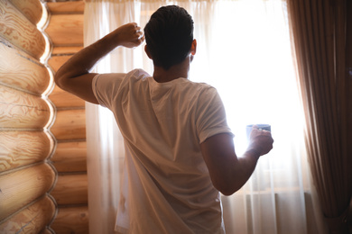 Man with drink stretching near window indoors. Lazy morning