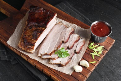 Photo of Pieces of baked pork belly served with sauce and parsley on black textured table, above view