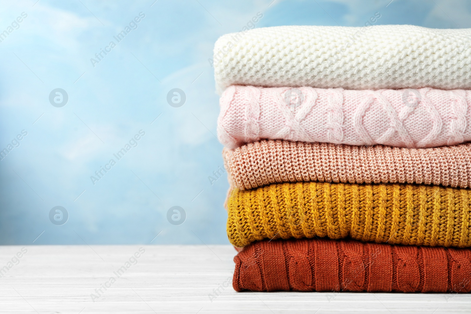 Photo of Stack of warm clothes on white wooden table against light blue background, space for text. Autumn season