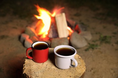 Photo of Cups of hot drink on old log near bonfire outdoors. Camping season