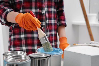Photo of Woman dipping brush into bucket of light blue paint indoors, closeup
