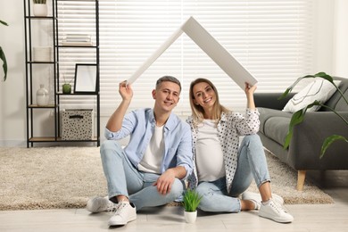 Young family housing concept. Pregnant woman with her husband sitting under cardboard roof on floor at home