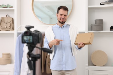 Photo of Smiling fashion blogger pointing at cardboard box while recording video at home