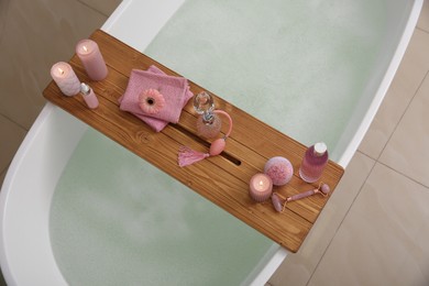 Photo of Wooden bath tray with candles and personal care products on tub indoors, above view