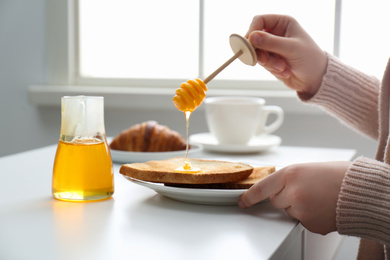 Woman pouring honey onto toast at white table, closeup