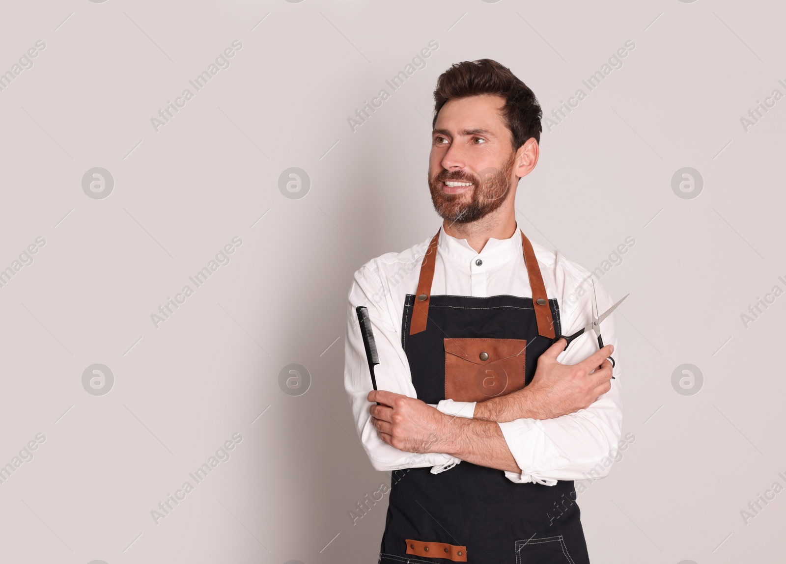 Photo of Smiling hairdresser in apron holding scissors and comb on light grey background, space for text