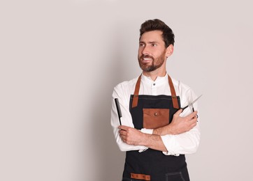 Smiling hairdresser in apron holding scissors and comb on light grey background, space for text