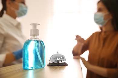 Receptionist with client at countertop in hotel, focus on dispenser bottle of antiseptic gel and service bell