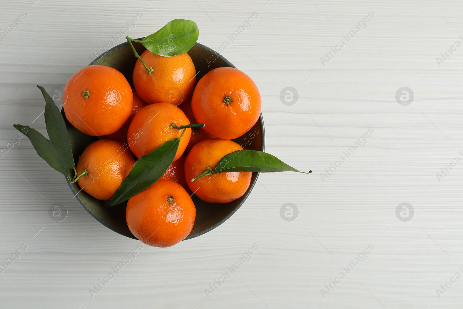 Photo of Delicious tangerines with green leaves in bowl on white wooden table, top view. Space for text